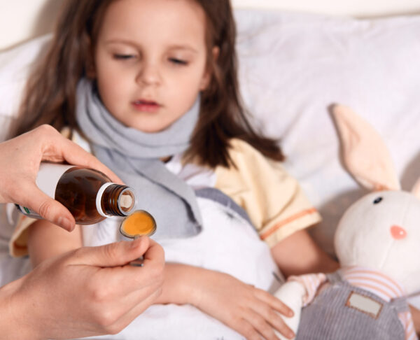 Horizontal photo of unknown hand pouring liquid into spoon from little bottle with syrup, person caring about child lying in bed with flu and sore throat, being upset. Children and health concept.